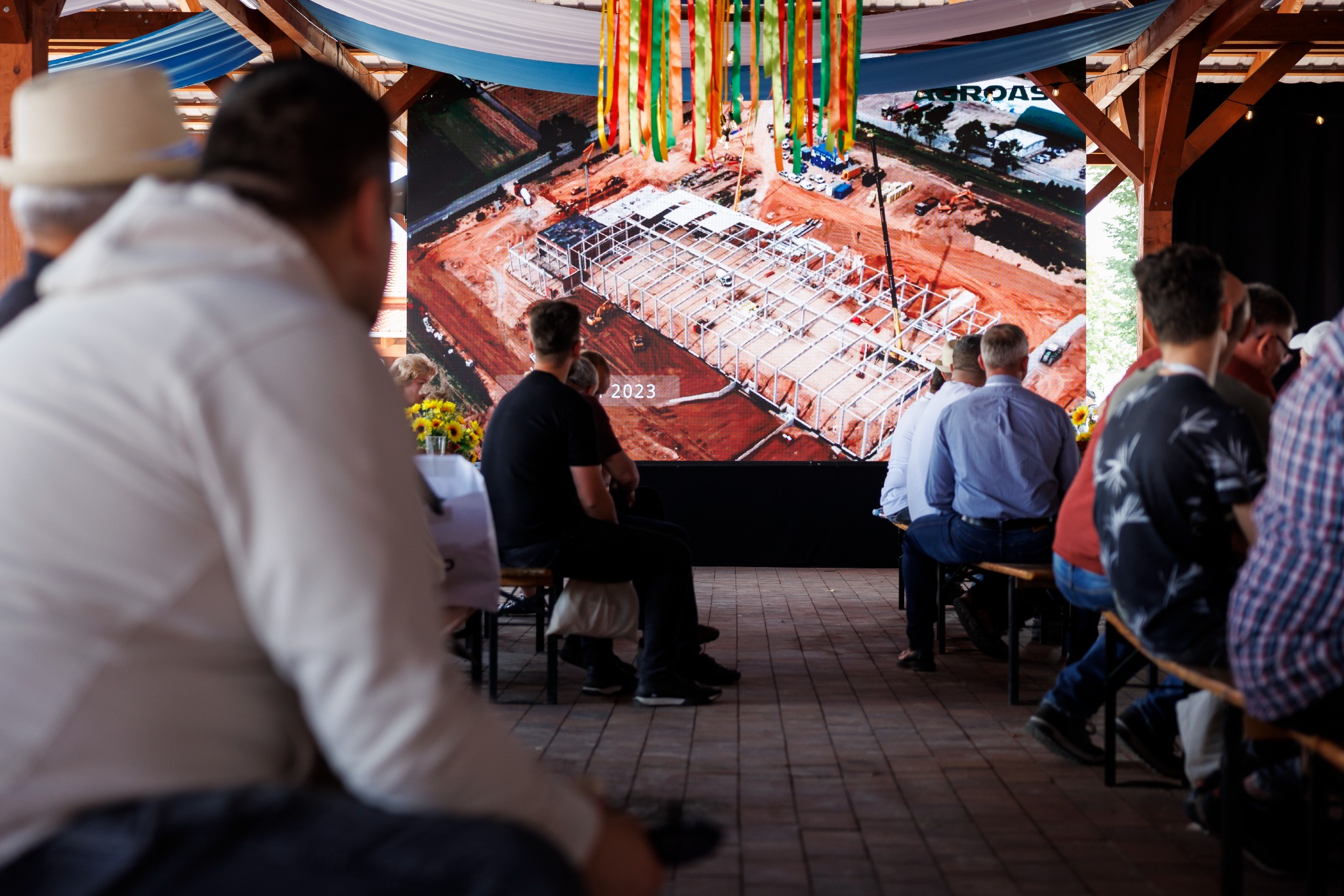 Widok z tyłu grupy ludzi siedzących na ławkach i oglądających prezentację na dużym ekranie przedstawiającym budowę hali przemysłowej z lotu ptaka, z dekoracjami z kolorowych wstążek i kwiatami.