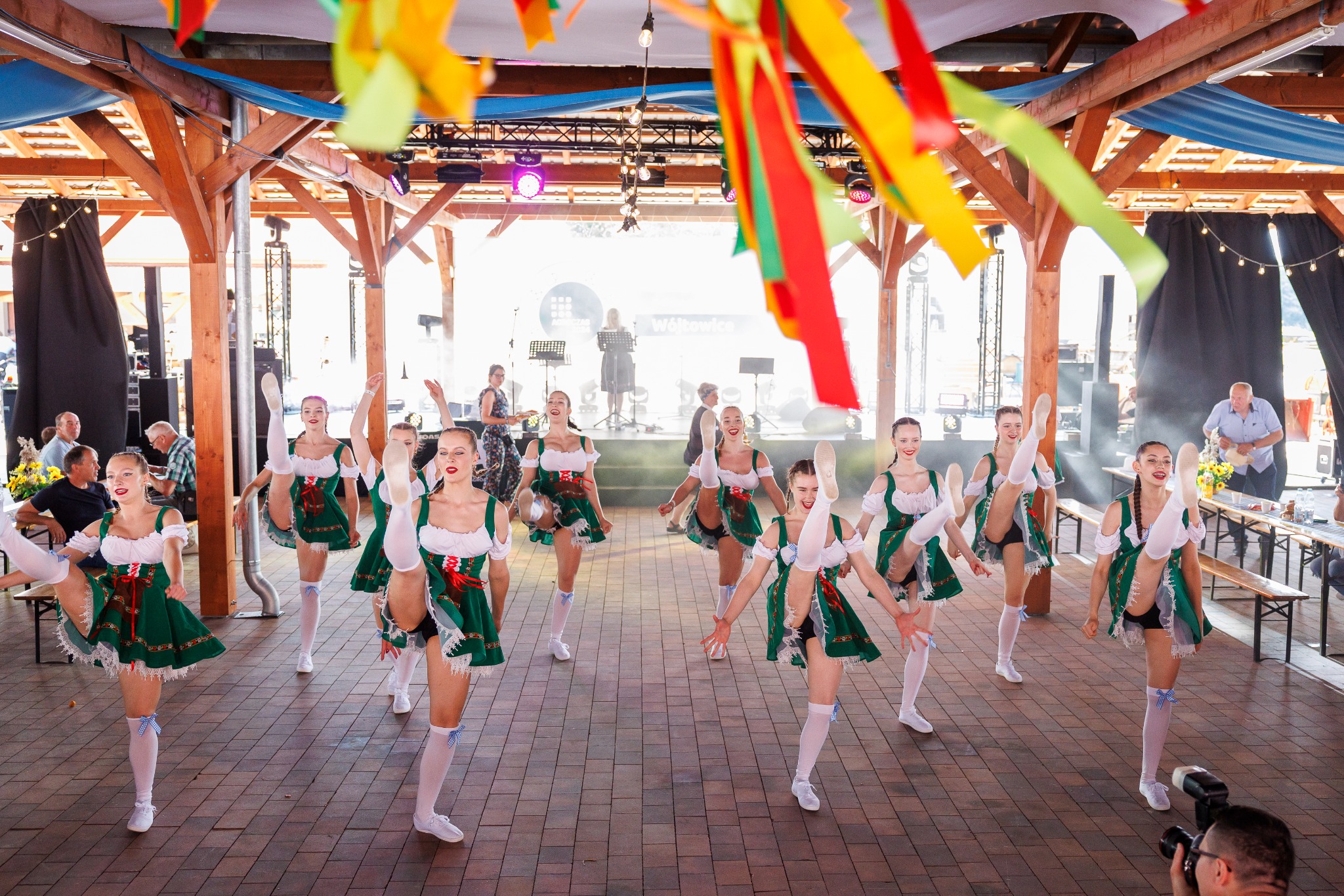 Grupa tancerek w bawarskich strojach wykonuje synchroniczny układ taneczny na scenie podczas imprezy plenerowej, z widoczną orkiestrą w tle.
