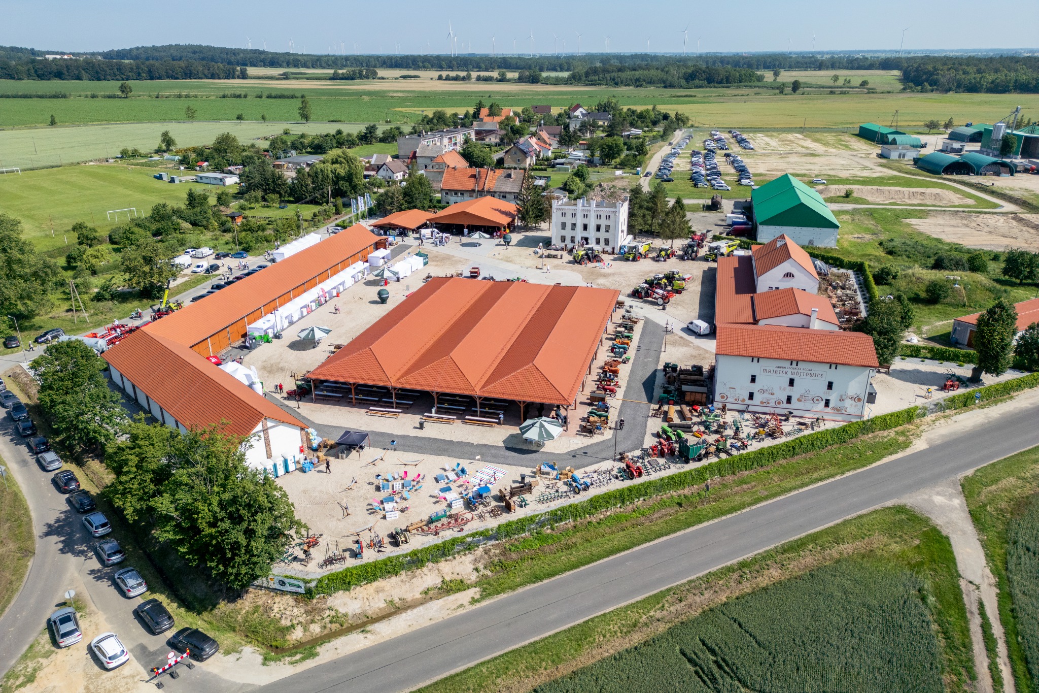 Widok z lotu ptaka na duży teren eventowy z czerwonymi dachami budynków, parkingiem, polami uprawnymi i turbinami wiatrowymi w oddali.