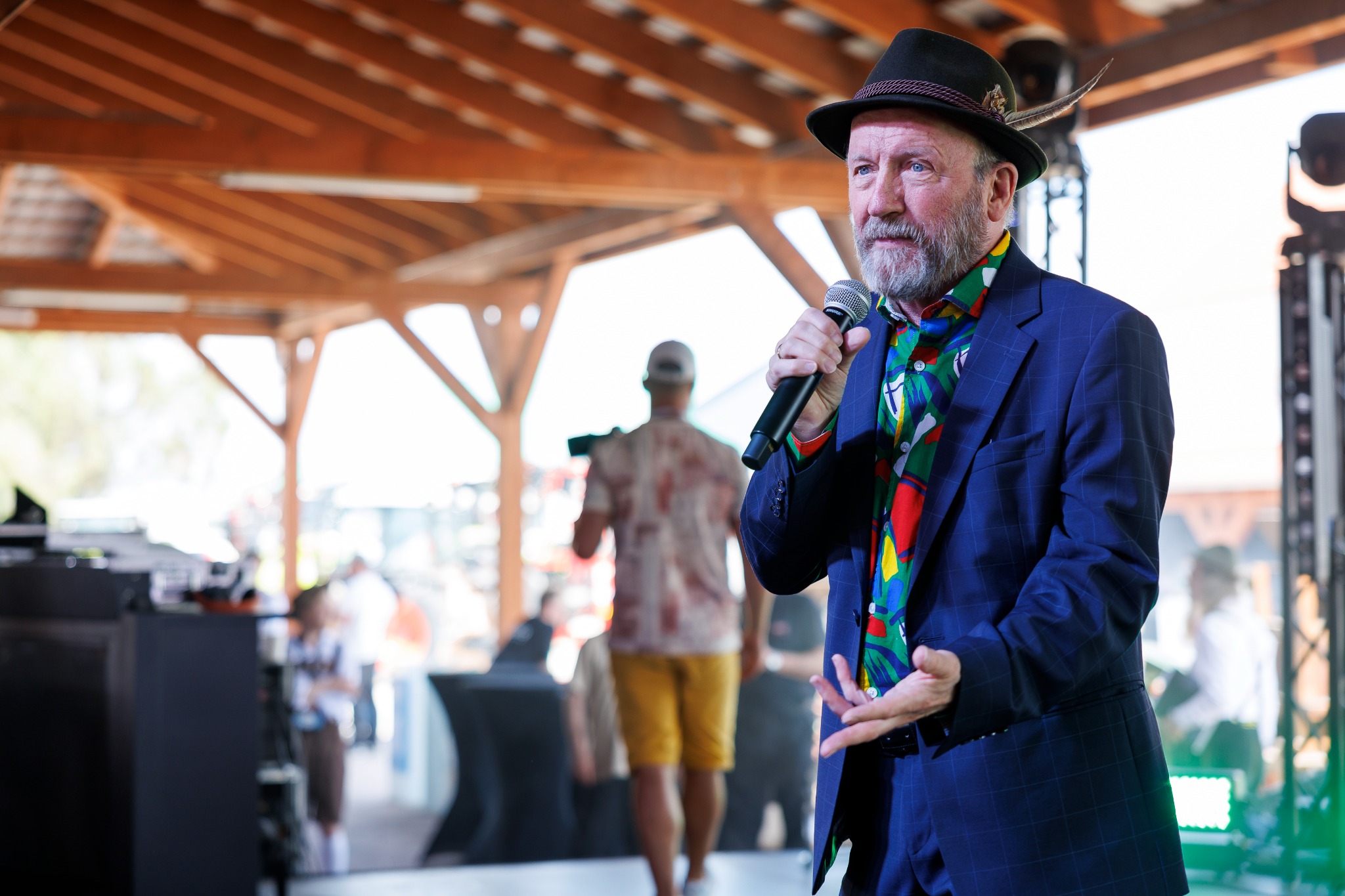 Mężczyzna z brodą w kapeluszu z piórem, w niebieskim garniturze i kolorowej koszuli, przemawia do mikrofonu na tle drewnianej konstrukcji i rozmytych postaci.