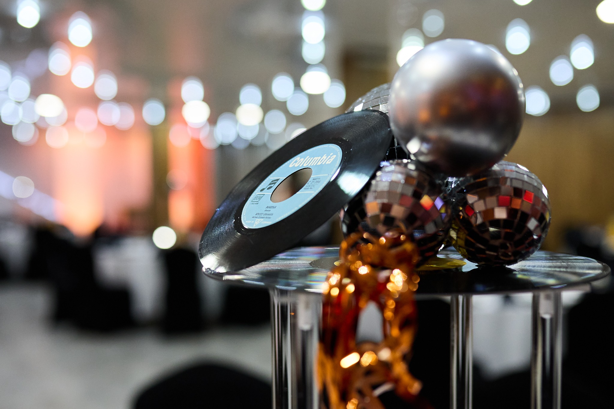 Dekoracja stołu z płytą winylową Columbia, srebrną kulą i małymi kulami dyskotekowymi na metalowym stojaku, w tle rozmyte światła i stoły nakryte na przyjęcie.