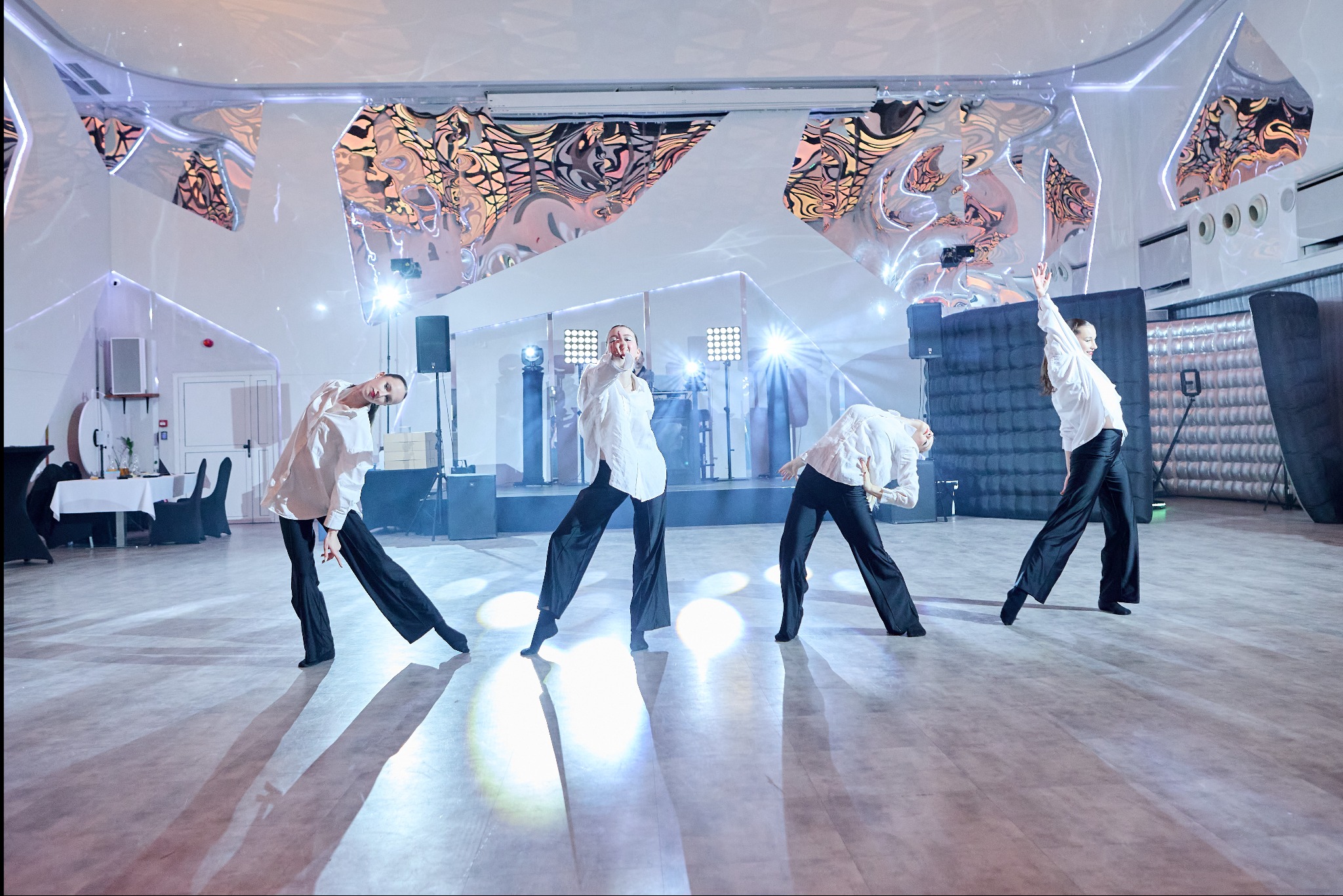 Cztery tancerki w białych koszulach i czarnych spodniach wykonują synchroniczny układ taneczny na scenie z futurystyczną dekoracją sufitu i reflektorami.