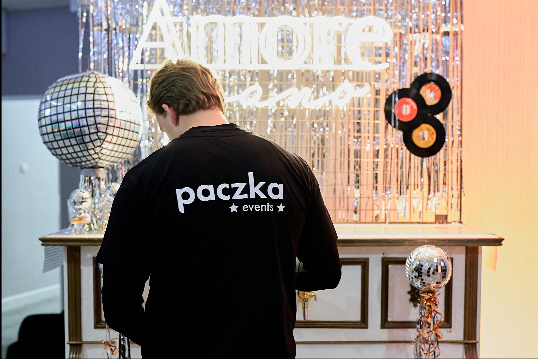 Osoba w czarnej koszulce z napisem 'paczka events' stoi tyłem do kamery przed dekoracją imprezową z neonowym napisem 'Amore', kulą dyskotekową i wiszącymi winylowymi płytami.