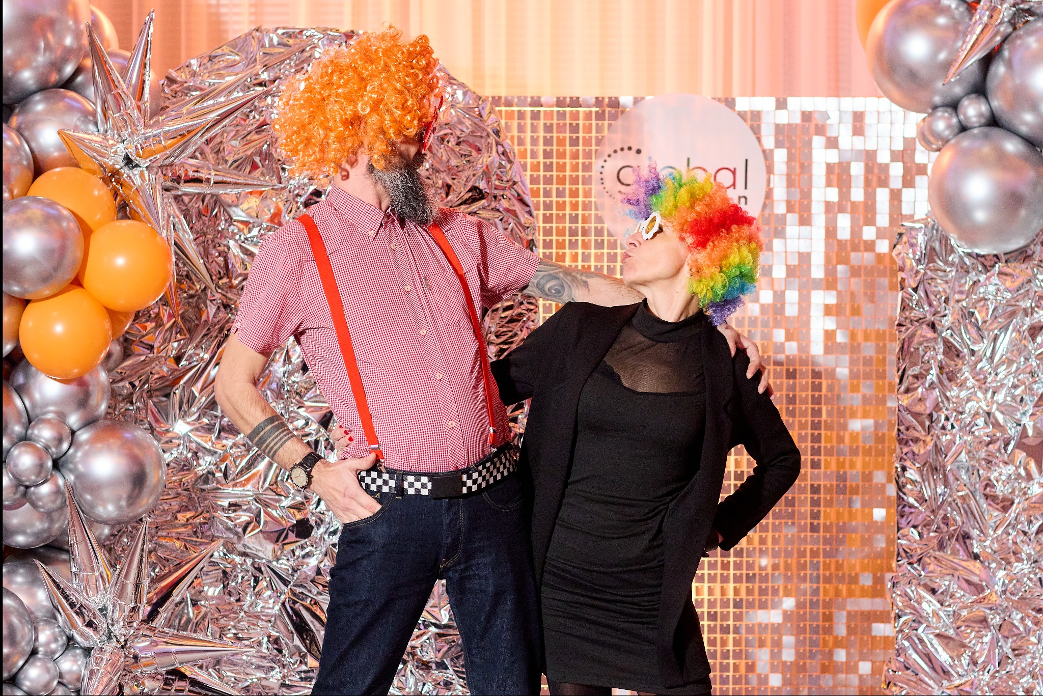 Para w kolorowych perukach, mężczyzna w czerwonej koszuli z szelkami, kobieta w czarnej sukience i marynarce, pozuje na tle srebrnej folii i balonów, stylizacja na imprezę tematyczną.