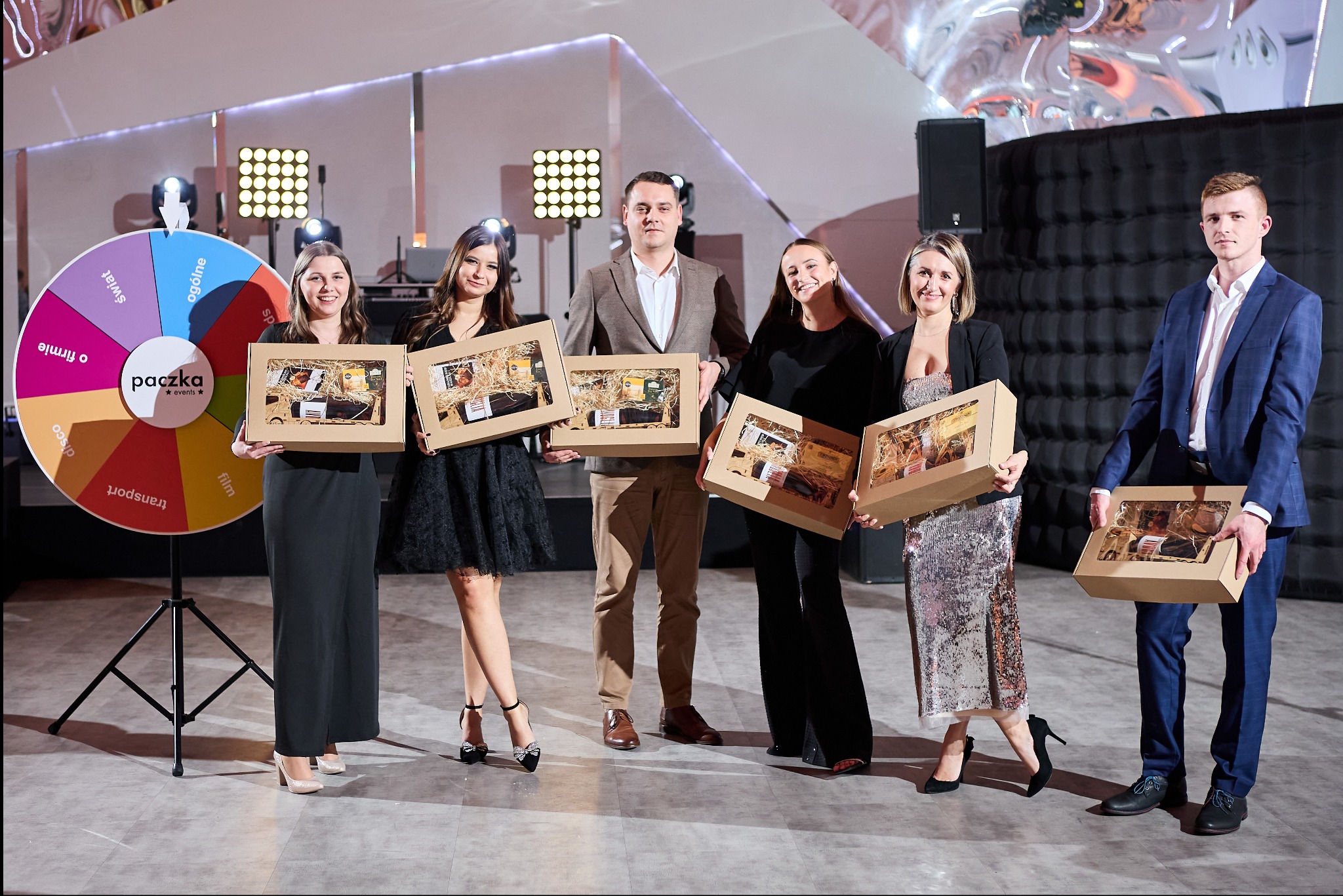 Grupa osób w eleganckich strojach prezentuje upominki w kartonowych pudełkach z okienkami, widoczne koło fortuny z napisem 'paczka events' w tle, oświetlenie sceniczne.