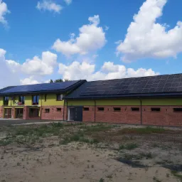 Dwukondygnacyjny budynek z panelami fotowoltaicznymi na dachu, wykonany z cegły i otynkowany na jasnozielono, na tle błękitnego nieba z białymi chmurami.