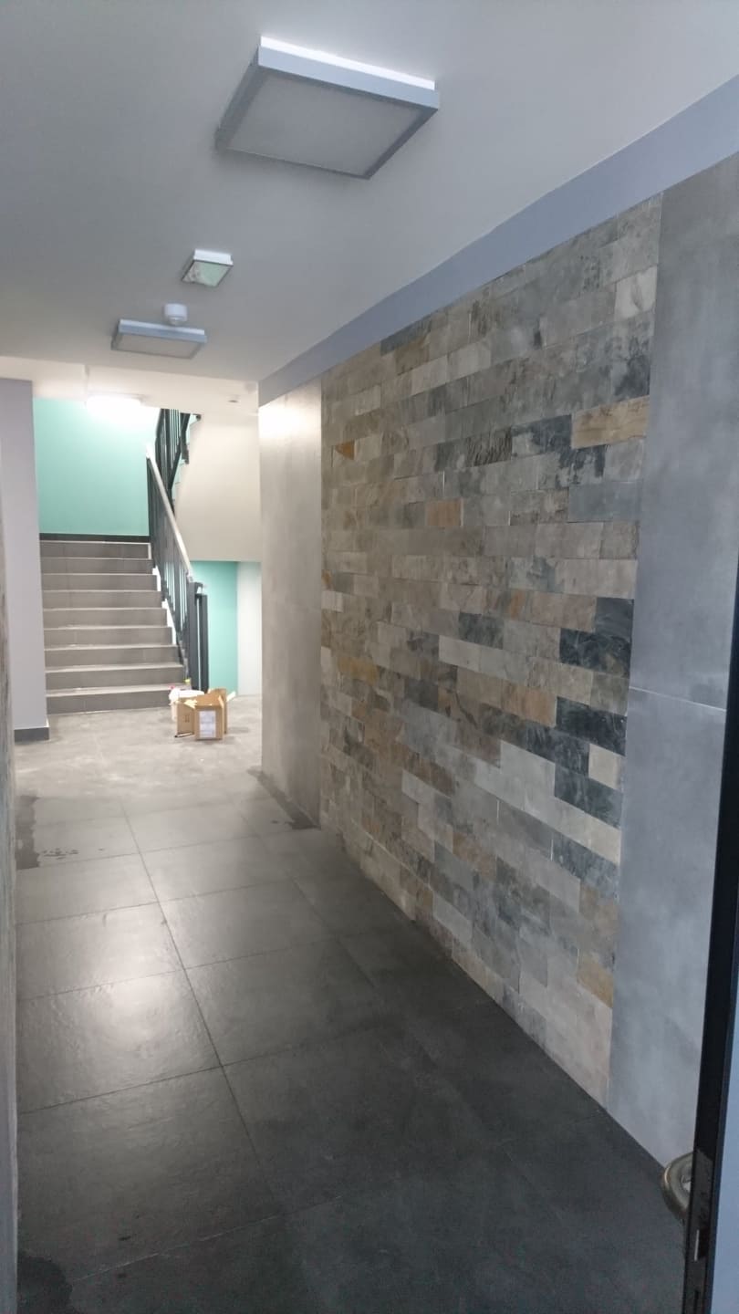 Korytarz z wyłożoną kamieniem dekoracyjnym ścianą, szara podłoga z płytek, schody w tle.