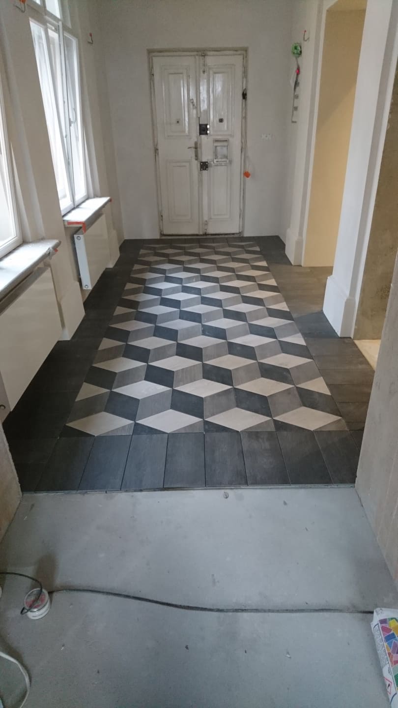 Korytarz z ułożoną podłogą z płytek w geometryczny wzór 3D, widoczne niedokończone prace wykończeniowe przy ścianach i podłodze.