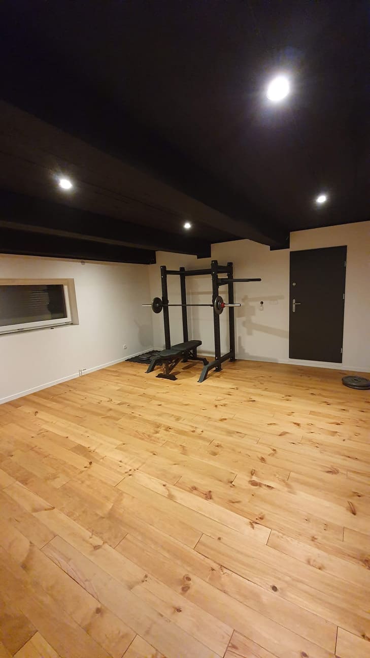 Jasna drewniana podłoga w domowej siłowni z czarnym sufitem, widoczne sztangi i ławeczka do ćwiczeń.