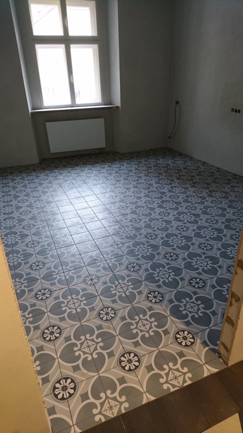 Ułożona podłoga z płytek w odcieniach szarości i błękitu, wzór geometryczno-kwiatowy, widoczne niedokończone wykończenie przy progu, surowe ściany i okno w tle.