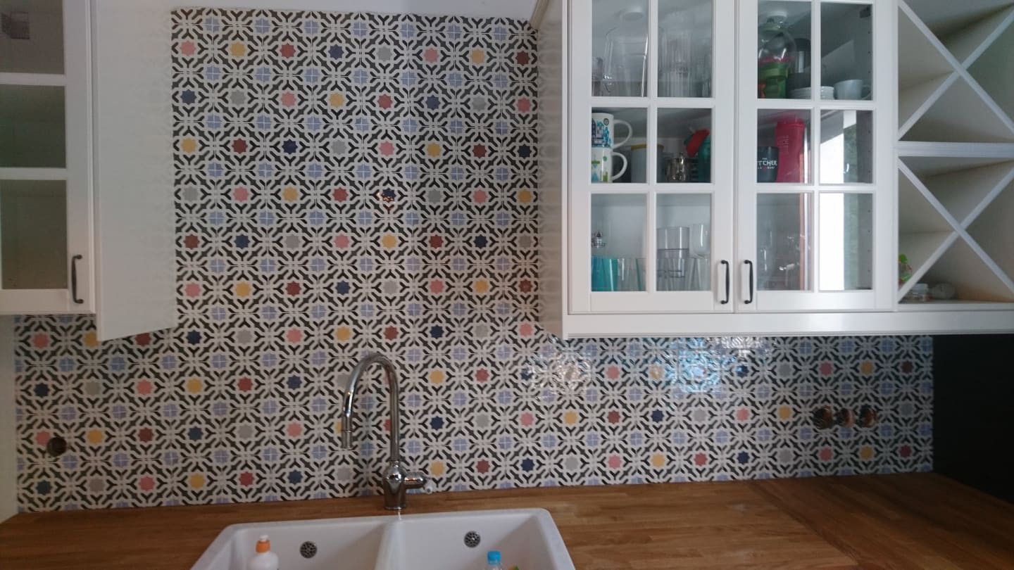 Wzorzyste płytki ceramiczne na ścianie nad drewnianym blatem kuchennym, z widocznym zlewem i baterią oraz białymi szafkami wiszącymi z przeszklonymi drzwiczkami.