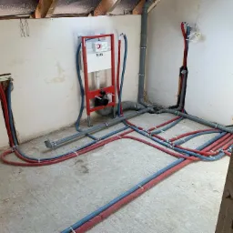 Surowy stan instalacji hydraulicznej w trakcie montażu: widoczne rury w izolacji czerwono-niebieskiej, szare rury kanalizacyjne oraz stelaż podtynkowy WC marki TECE zamontowany na ścianie.