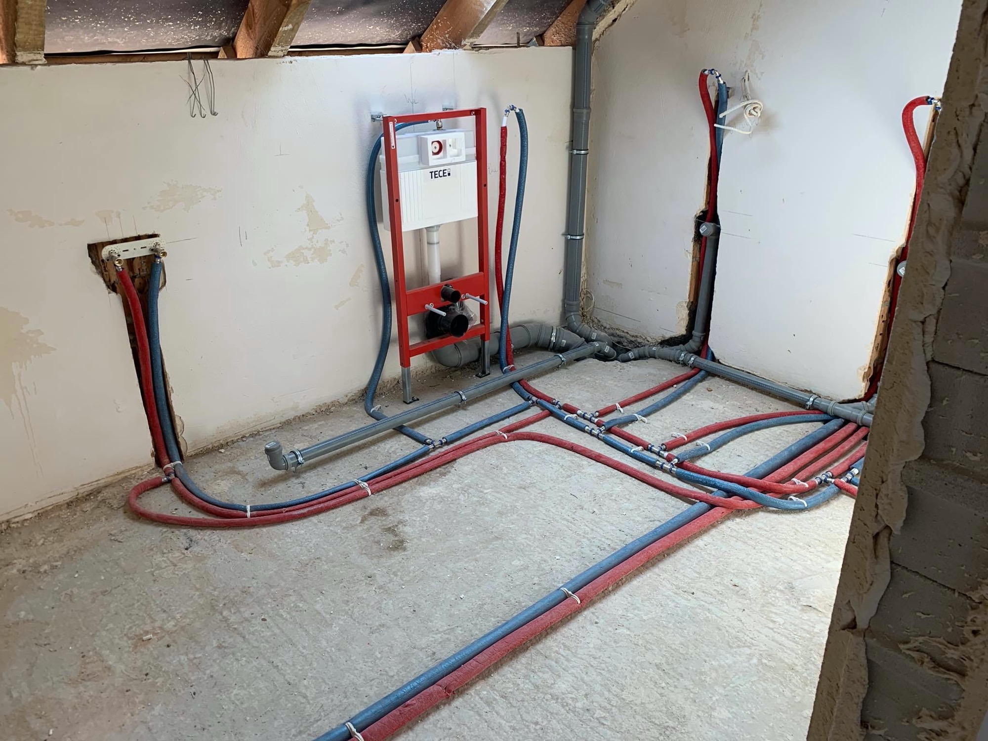 Surowy stan instalacji hydraulicznej w trakcie montażu: widoczne rury w izolacji czerwono-niebieskiej, szare rury kanalizacyjne oraz stelaż podtynkowy WC marki TECE zamontowany na ścianie.