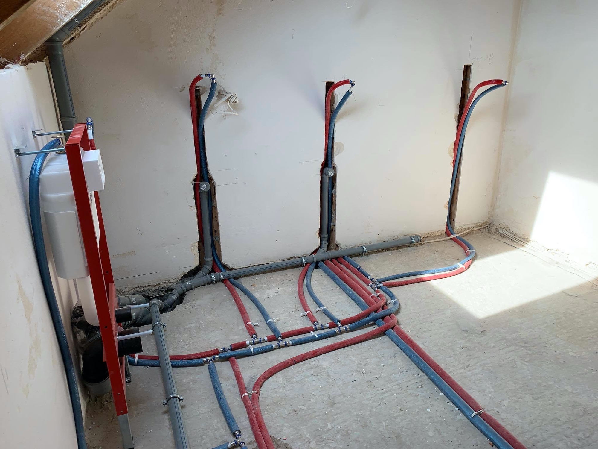 Rozbudowana instalacja wodna i kanalizacyjna w surowym pomieszczeniu, widoczne rury PEX w izolacji czerwonej i niebieskiej, pion kanalizacyjny, stelaż podtynkowy WC z widoczną spłuczką.