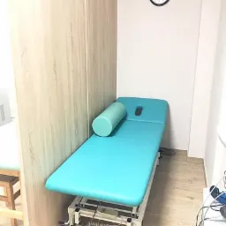 Turkusowy stół do zabiegów z wałkiem, ustawiony w rogu jasnego gabinetu z zegarem na ścianie.