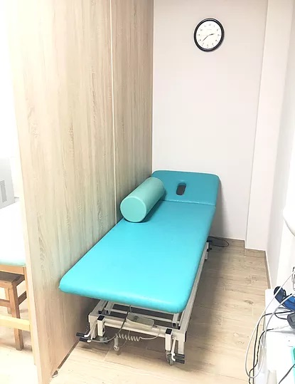 Turkusowy stół do zabiegów z wałkiem, ustawiony w rogu jasnego gabinetu z zegarem na ścianie.