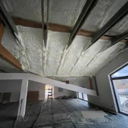 Poddasze po ociepleniu pianką PUR, widoczne drewniane belki konstrukcyjne, czarna folia ochronna na podłodze, drabina i okno z widokiem na zewnątrz.