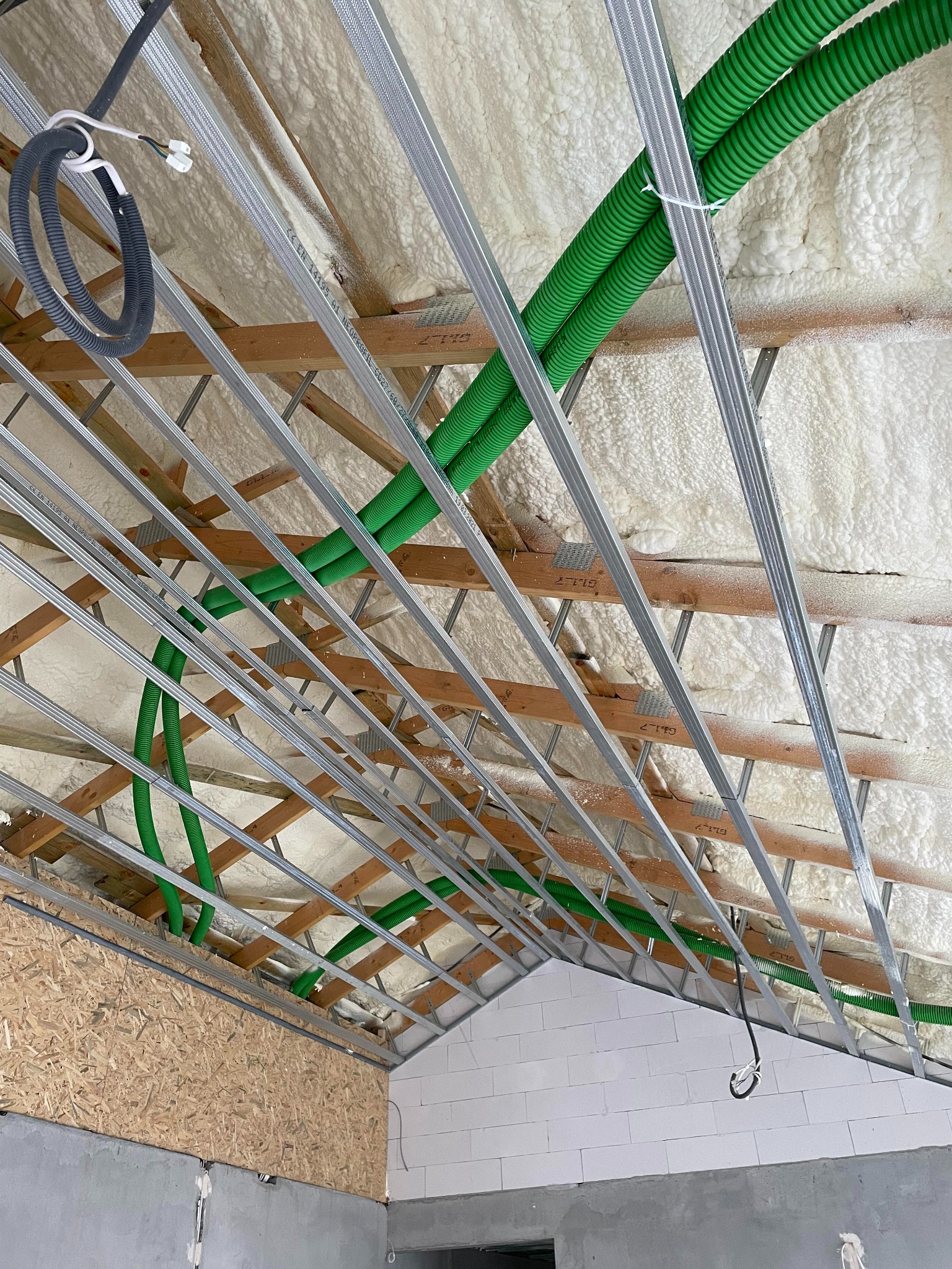 Poddasze w trakcie izolacji: natrysk piany PUR na drewnianą konstrukcję dachu, widoczne zielone rury instalacyjne i metalowe profile pod płyty gipsowo-kartonowe.