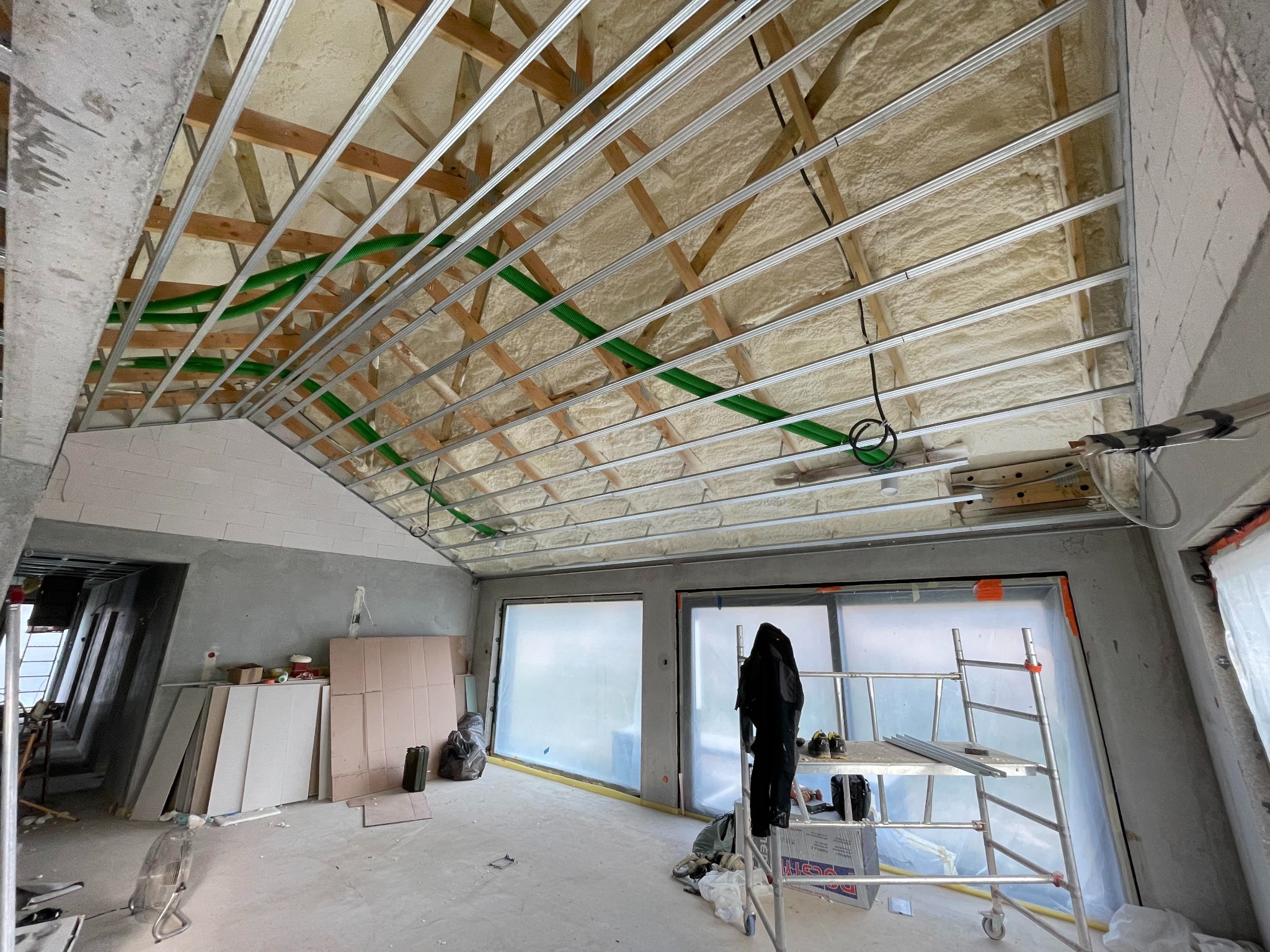 Poddasze w trakcie izolacji pianą, widoczne drewniane krokwie, metalowe profile, zielone rury instalacyjne i warstwa piany PUR, okna zabezpieczone folią ochronną, rusztowanie w pomieszczeniu.