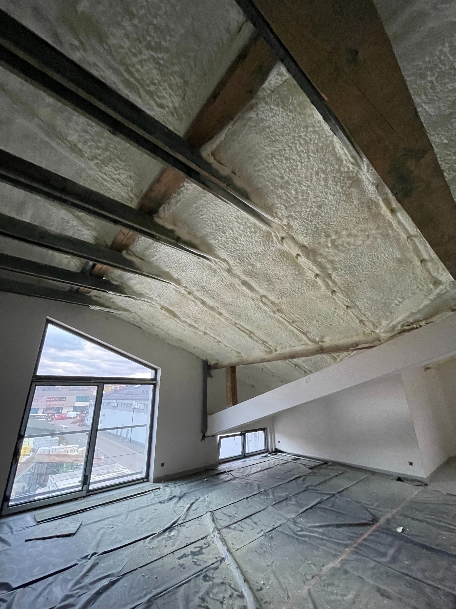 Poddasze po ociepleniu pianką PUR, widoczne drewniane i metalowe elementy konstrukcyjne dachu oraz okno o nietypowym, trójkątnym kształcie. Podłoga zabezpieczona folią malarską.