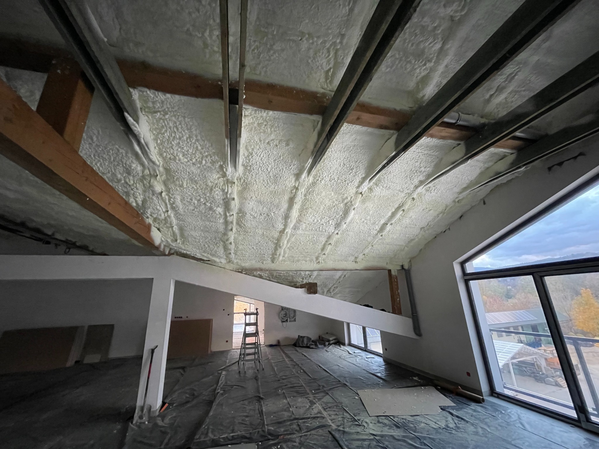 Poddasze po ociepleniu pianką PUR, widoczne drewniane belki konstrukcyjne, czarna folia ochronna na podłodze, drabina i okno z widokiem na zewnątrz.