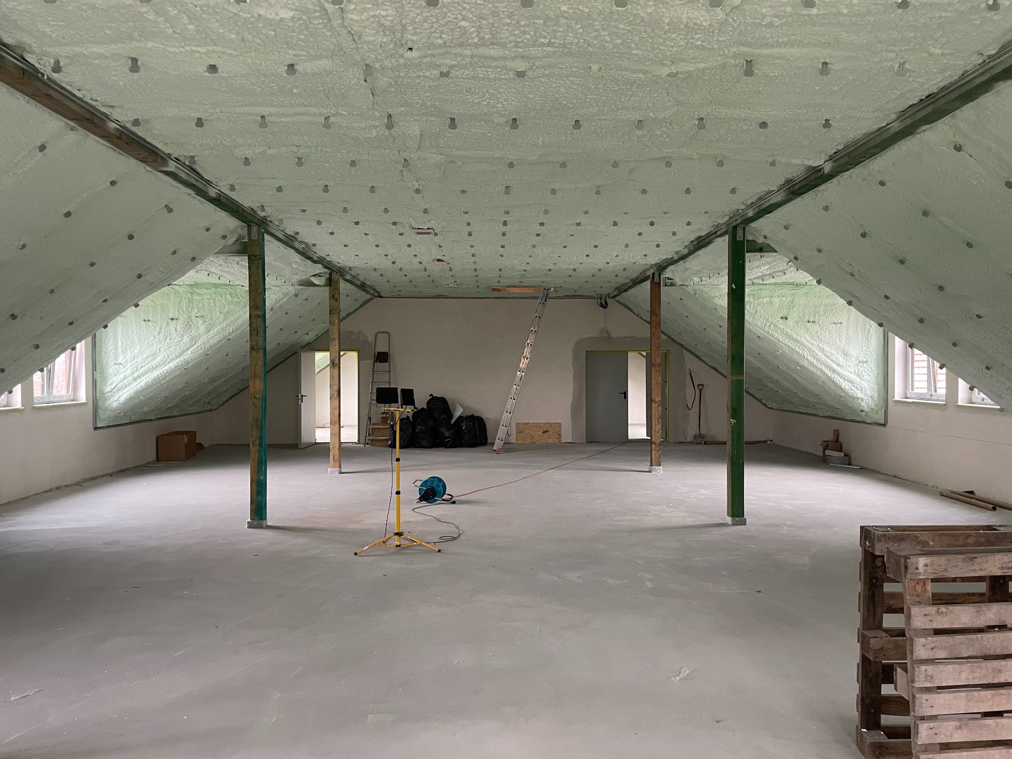 Poddasze po izolacji pianą, widoczne krokwie i równomiernie nałożona warstwa piany PUR z widocznymi punktami mocowania, na podłodze widoczny sprzęt budowlany i drabina oparta o ścianę.