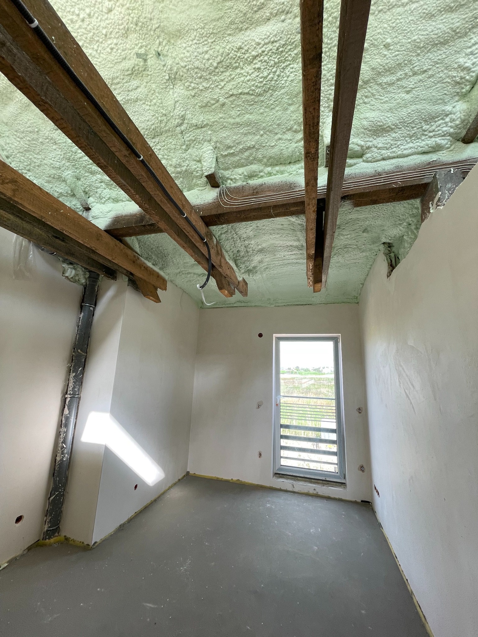 Poddasze po izolacji pianką PUR, widoczne drewniane belki stropowe, instalacje elektryczne i rury kanalizacyjne, ściany przygotowane do wykończenia, okno z widokiem na zewnątrz.