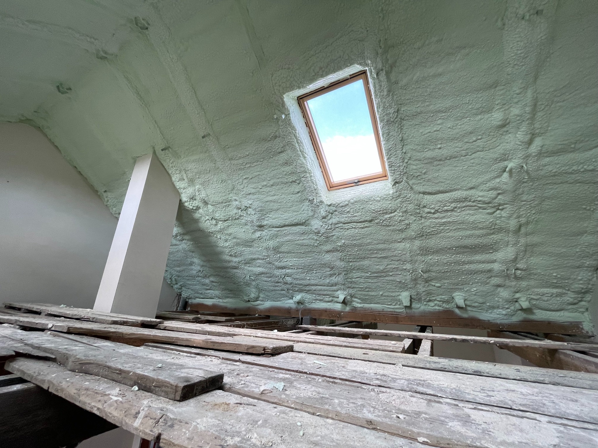 Poddasze po ociepleniu pianką PUR, widoczne deski podłogowe na pierwszym planie i okno dachowe w tle, piana jasna, równomiernie nałożona.