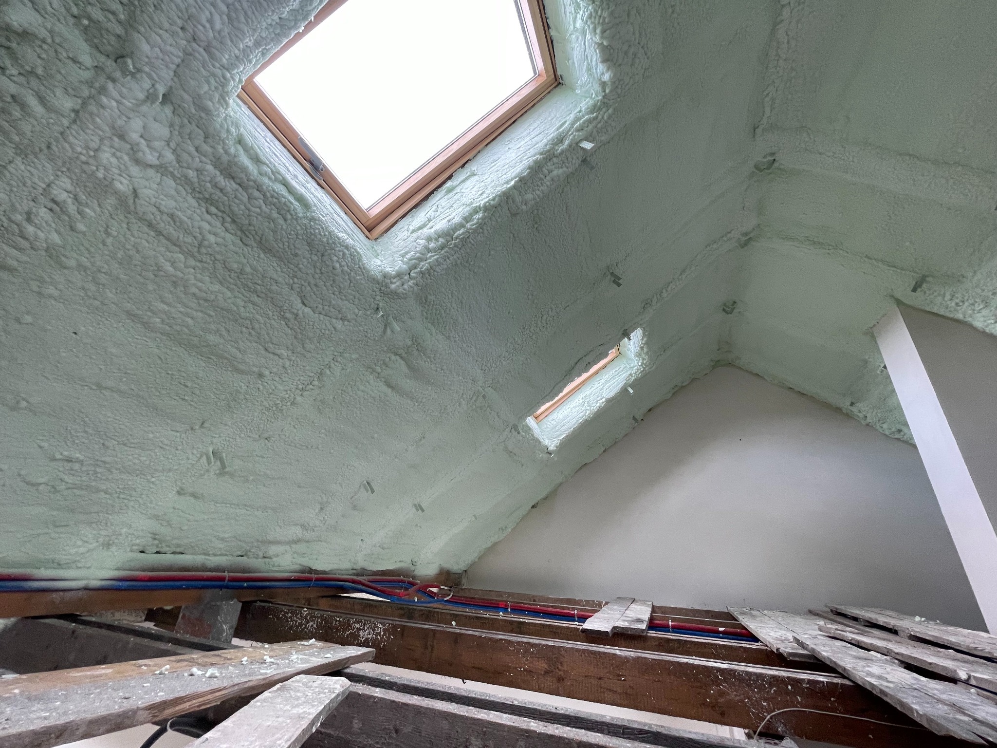 Poddasze po izolacji pianą, widoczne okna dachowe i instalacja wodna na drewnianej konstrukcji stropu.