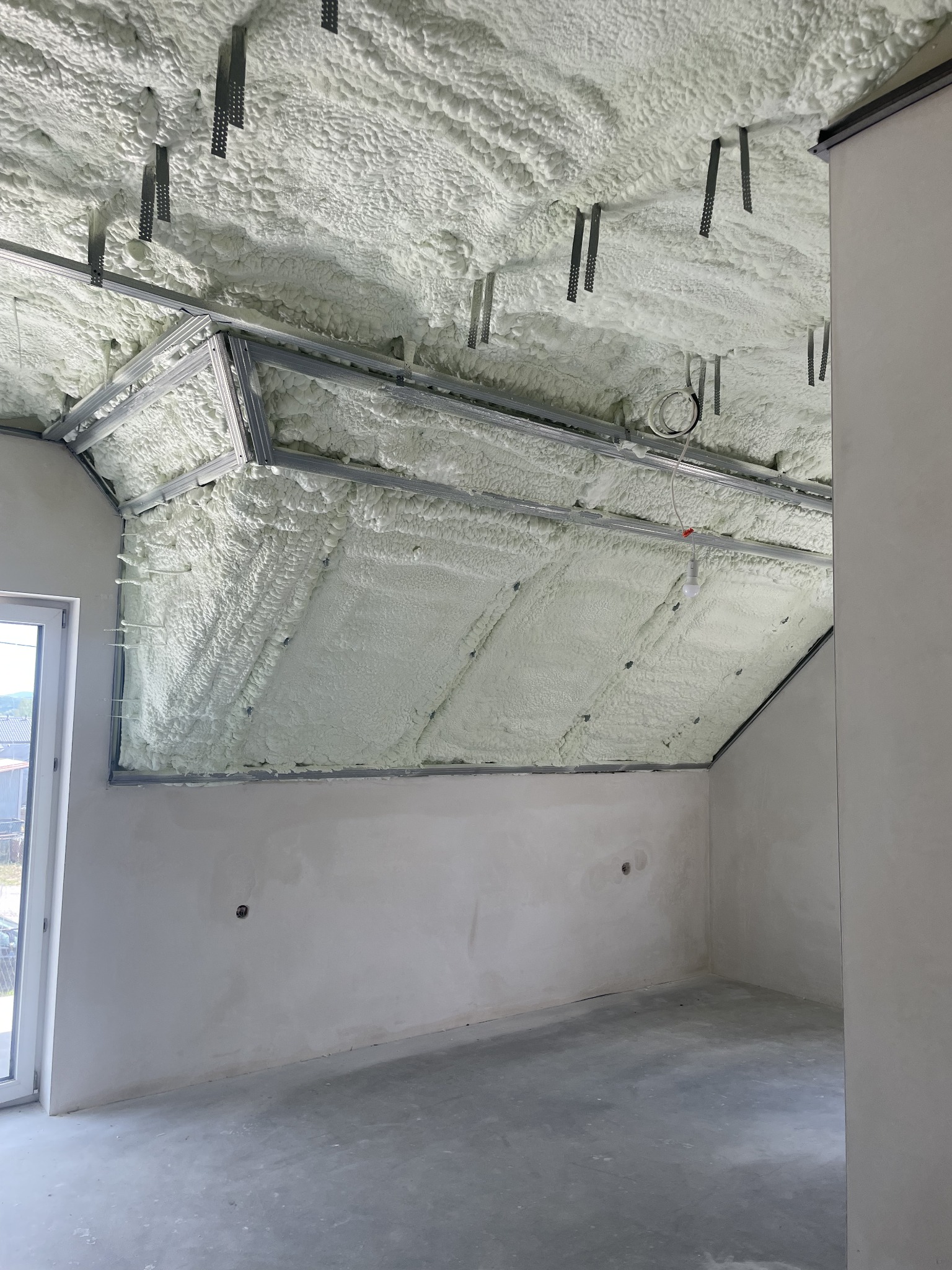 Poddasze w trakcie izolacji pianą, widoczne metalowe profile konstrukcyjne i świeżo nałożona, nierówna warstwa piany PUR, zwisające uchwyty montażowe, surowy stan wykończenia ścian i podłogi.