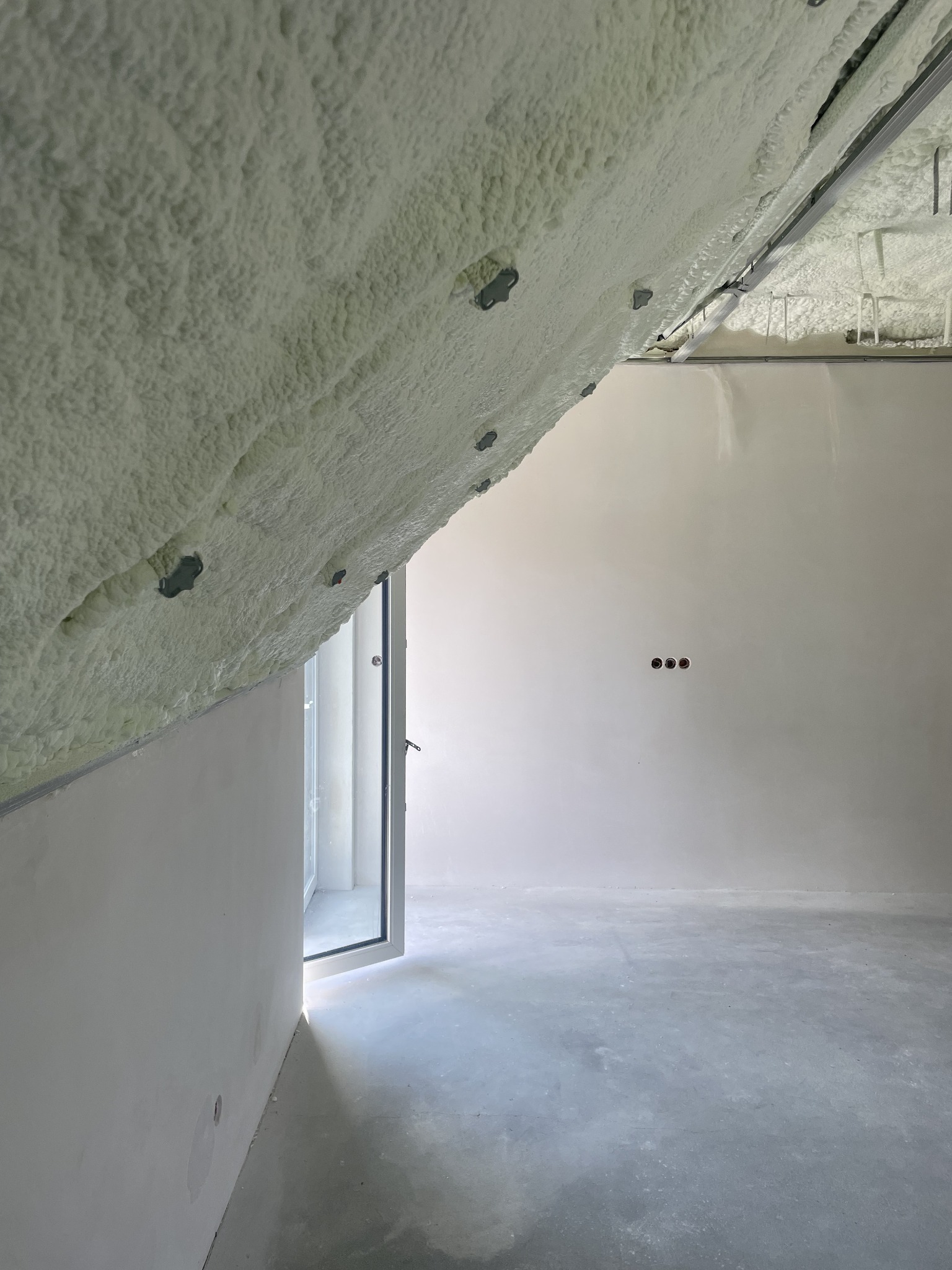 Poddasze po ociepleniu pianką PUR, widoczne metalowe elementy konstrukcyjne, surowa ściana i podłoga, otwarte drzwi balkonowe wpuszczające światło.