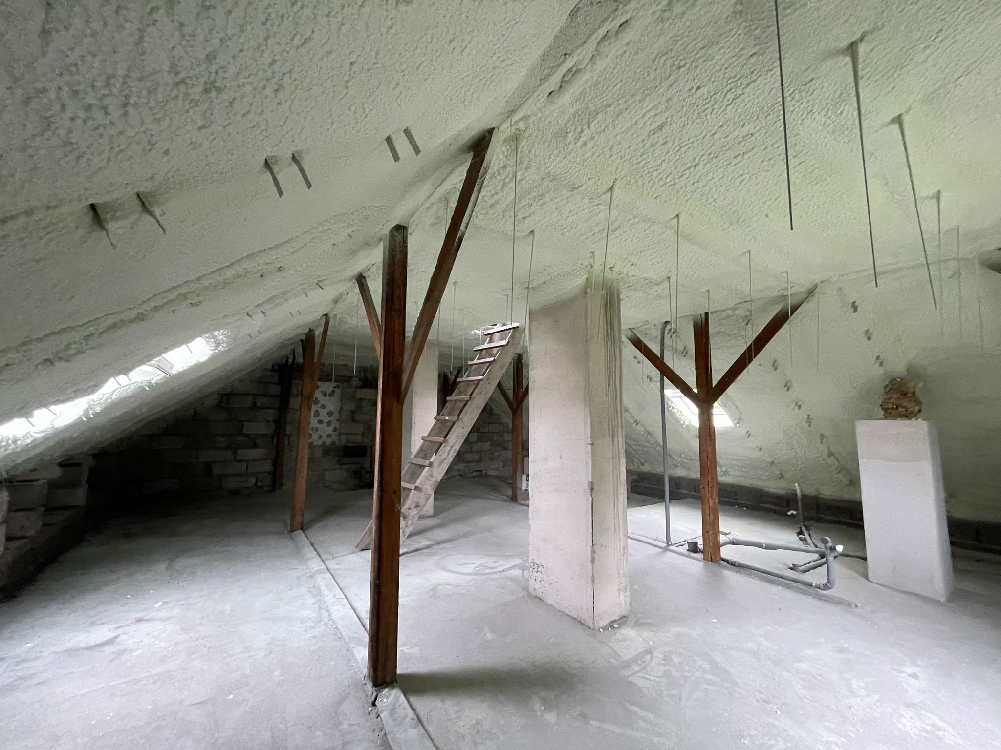 Poddasze po ociepleniu pianką: widoczne drewniane elementy konstrukcyjne, schody drabinkowe, wystające metalowe pręty oraz równomiernie nałożona warstwa izolacji piankowej na skośnym dachu.