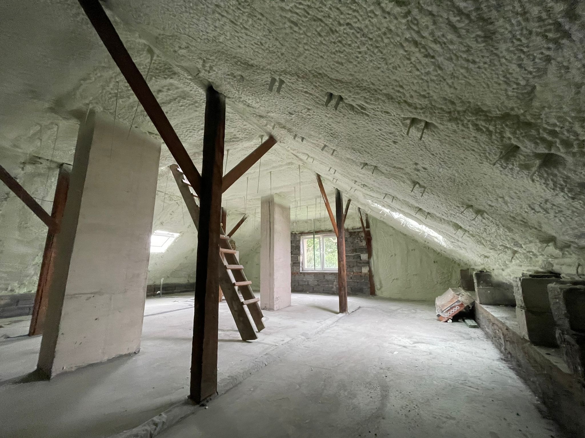 Poddasze po izolacji pianą, widoczne krokwie, słupy i drabina, ściany i sufit pokryte jasną pianą, okno dachowe i zwykłe okno w tle.