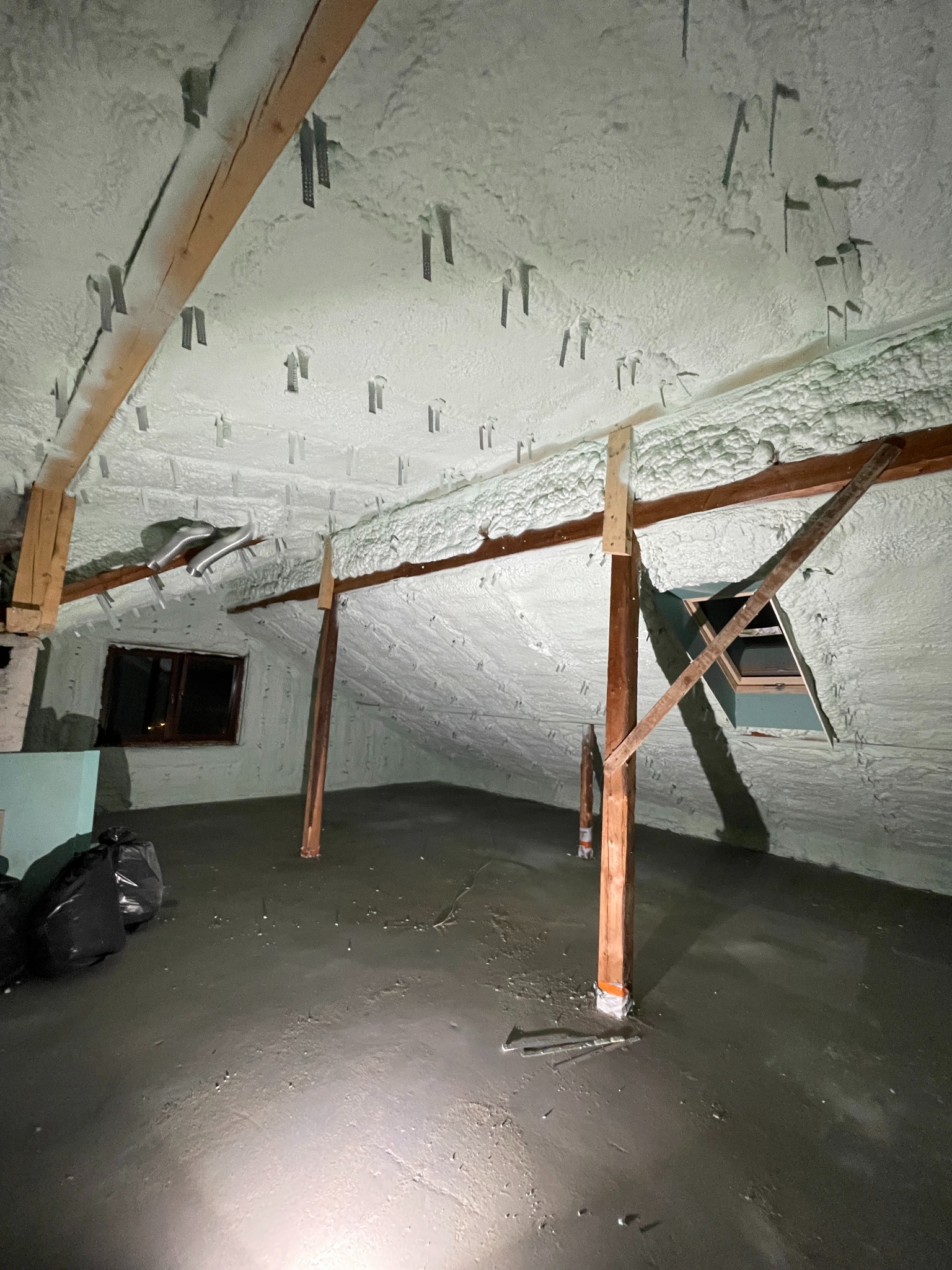 Poddasze po ociepleniu pianką PUR, widoczne krokwie i metalowe uchwyty mocujące, worki na śmieci w rogu pomieszczenia, surowe wykończenie podłogi.