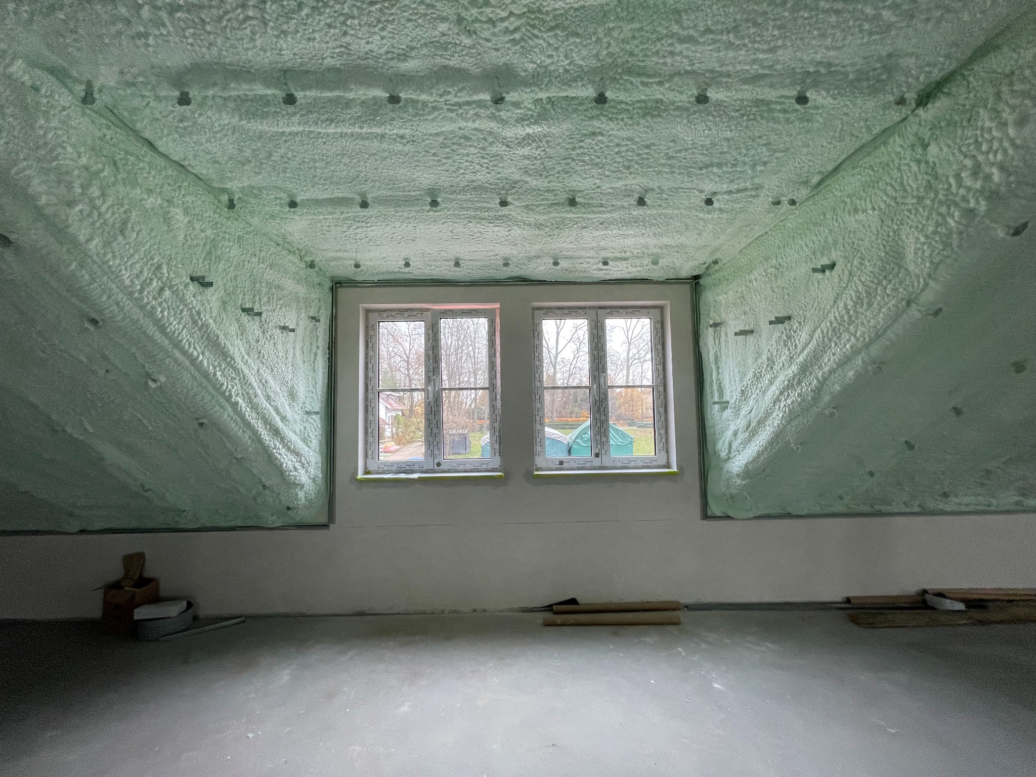 Poddasze po ociepleniu pianką PUR, widoczne dwa okna dachowe, szara podłoga i ściany, pianka o strukturze gąbki pokrywa skosy dachu z widocznymi mocowaniami.