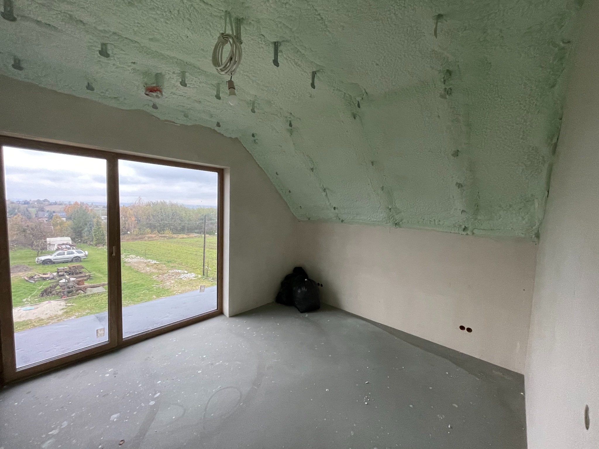 Poddasze po ociepleniu pianką PUR, widoczne świeżo nałożona warstwa pianki na skosach dachu, wystające kable z żarówką, okno z widokiem na zewnątrz, na podłodze leżą czarne worki.