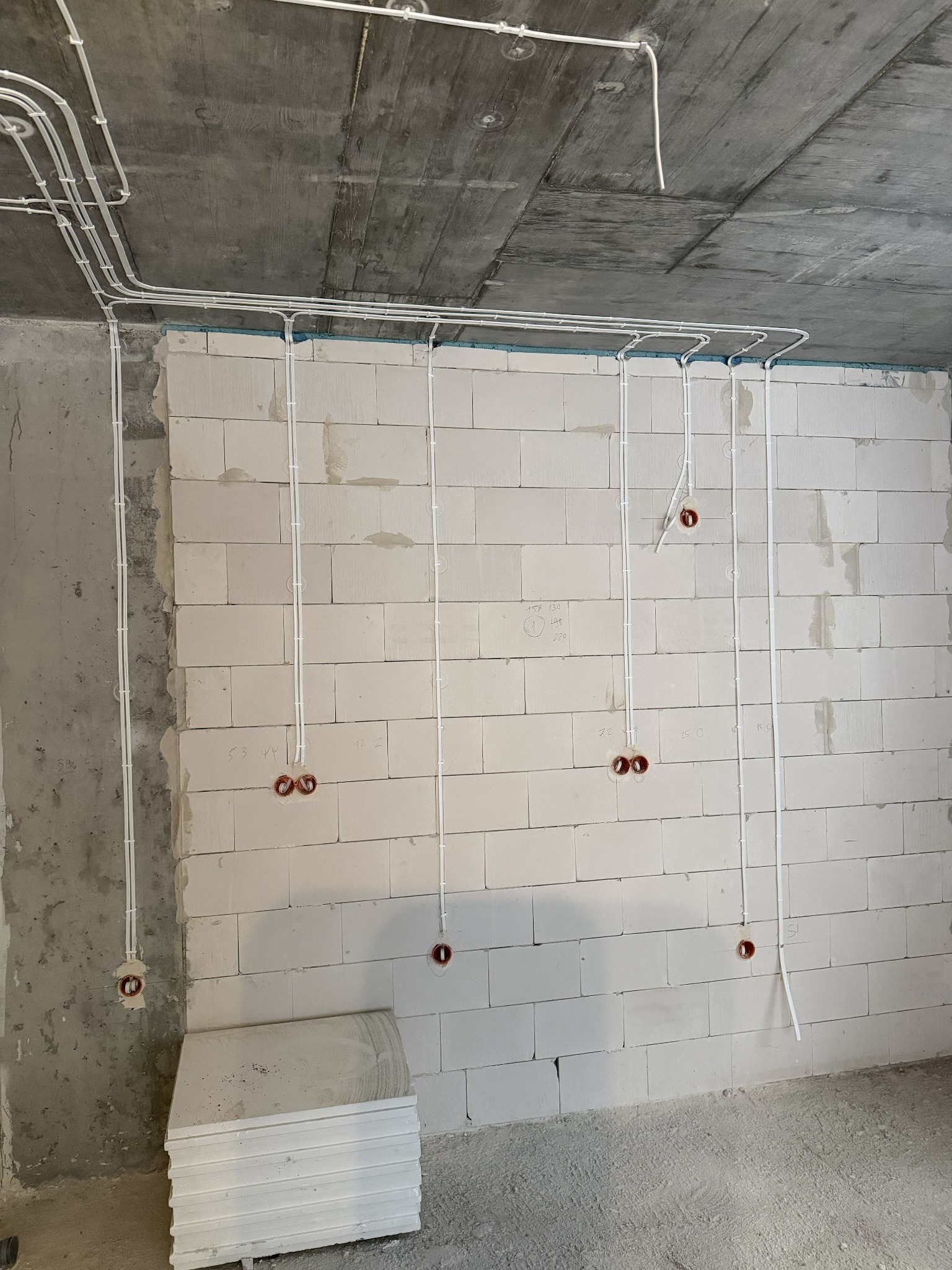 Białe rurki instalacji elektrycznej przymocowane do surowej ściany z betonu komórkowego, z widocznymi puszkami pod gniazdka. Etap prac instalacyjnych.