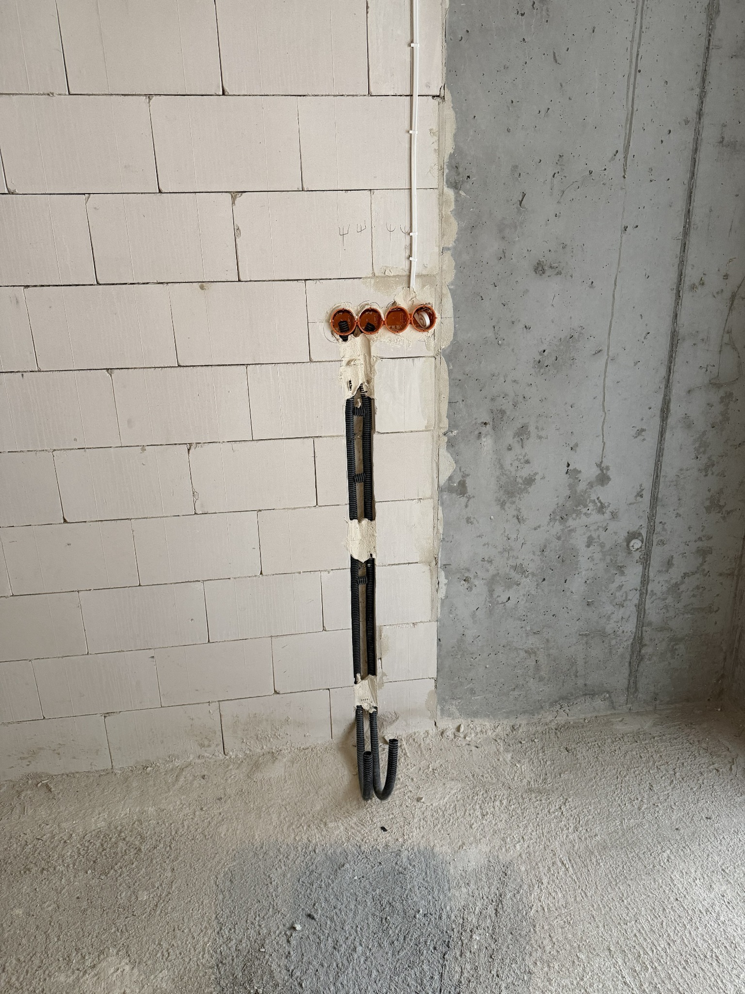 Surowa ściana z betonu i bloczków, z widocznymi puszkami instalacyjnymi i peszlami. Instalacja elektryczna w trakcie montażu, widoczne przewody.