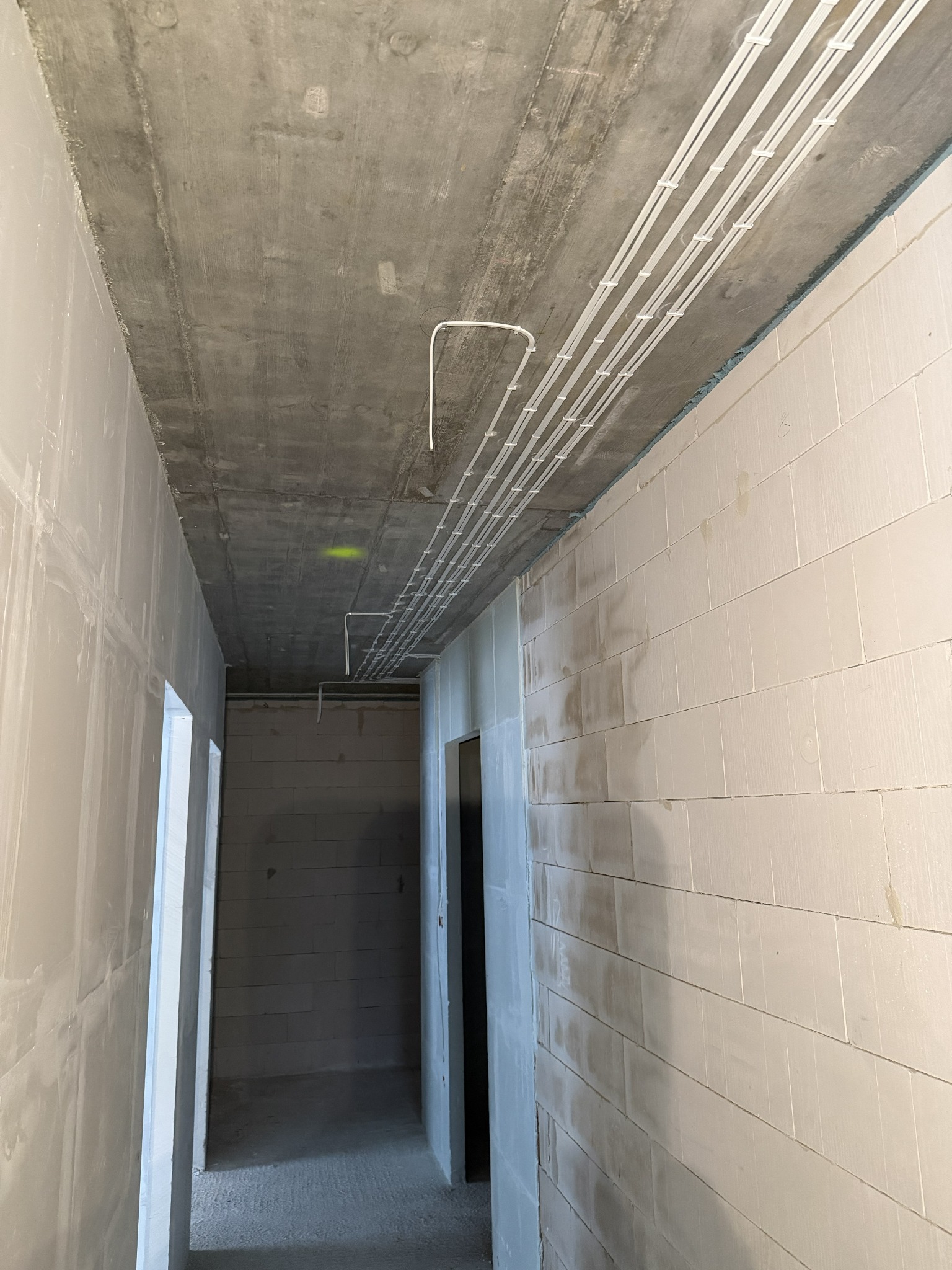 Białe przewody elektryczne zamocowane do surowego betonowego sufitu w korytarzu z białymi ścianami z bloków. Widoczne otwory drzwiowe.