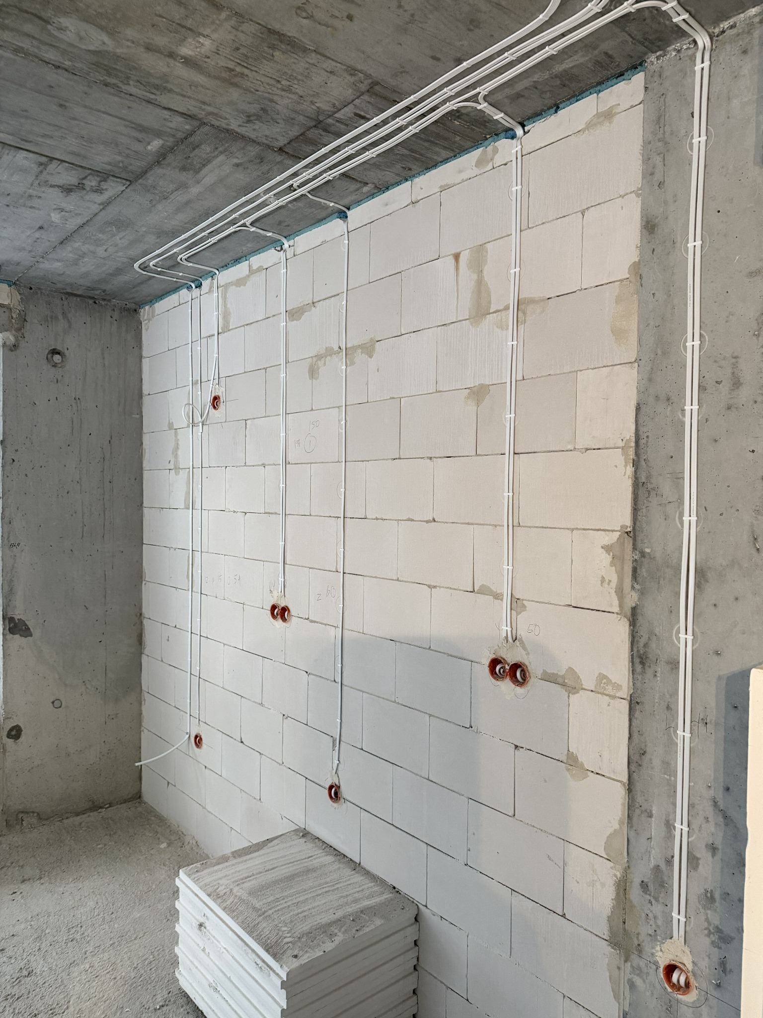 Białe kable elektryczne prowadzone po ścianie z betonu komórkowego i suficie w surowym stanie. Widoczne puszki instalacyjne. Instalacja elektryczna w trakcie realizacji.