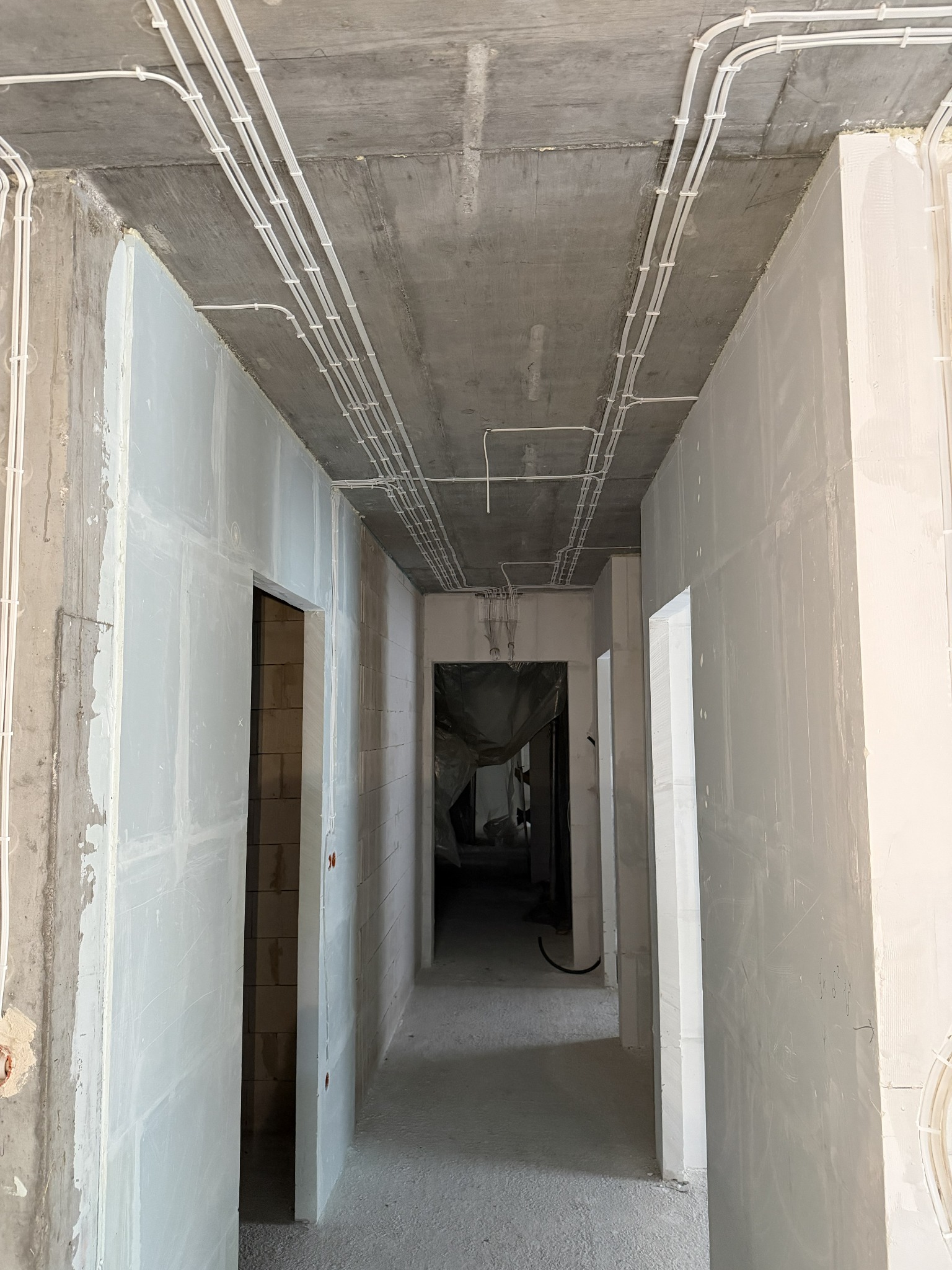 Korytarz w surowym stanie z widocznymi, poprowadzonymi po suficie przewodami elektrycznymi w białych peszlach, zamocowanymi uchwytami. Widoczne surowe ściany.