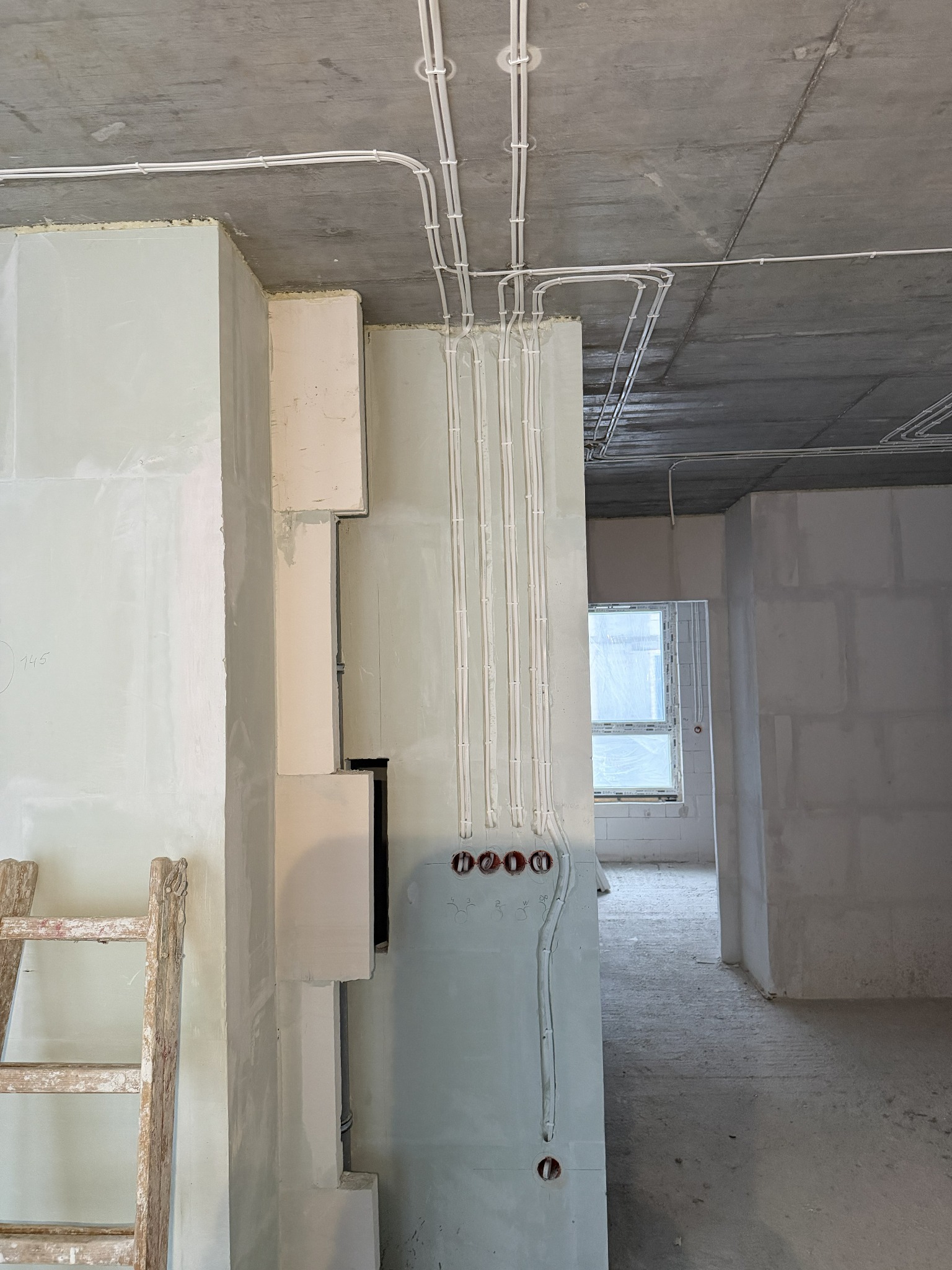 Białe kable elektryczne prowadzone po suficie i ścianie w surowym wnętrzu. Widoczne puszki instalacyjne i fragment drewnianej drabiny. Stan surowy, przed wykończeniem.