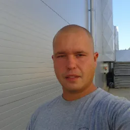 Mężczyzna na tle budynku z szarej blachy trapezowej z elementami wentylacji. Widoczny fragment trawnika i drzewa w tle. Dzień słoneczny, ostre światło.