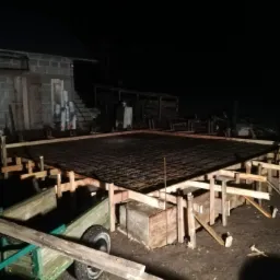 Przygotowanie pod fundament: drewniane szalunki zbrojone stalową siatką w nocnym oświetleniu, widoczny budynek gospodarczy w tle.