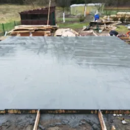 Świeżo wylana, gładka płyta betonowa w drewnianej szalunku, teren budowy w tle.