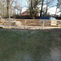 Nowo postawione, drewniane ogrodzenie panelowe z metalowymi słupkami na betonowej podmurówce, częściowo zasłaniające dwa zaparkowane samochody na tle drzew i domu z czerwoną dachówką.