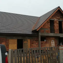 Dom w stanie surowym z częściowo położonym ciemnobrązowym dachem z dachówki ceramicznej, widoczna drewniana więźba dachowa, okna i drzwi zabite płytami OSB, w tle szary samochód dostawczy.