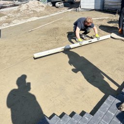 USŁUGI BUDOWLANE Damian Uzdowski - Pracownik w żółtych rękawicach wyrównuje piasek pod kostkę brukową za pomocą poziomicy; w tle mini koparka i palety z kostką.