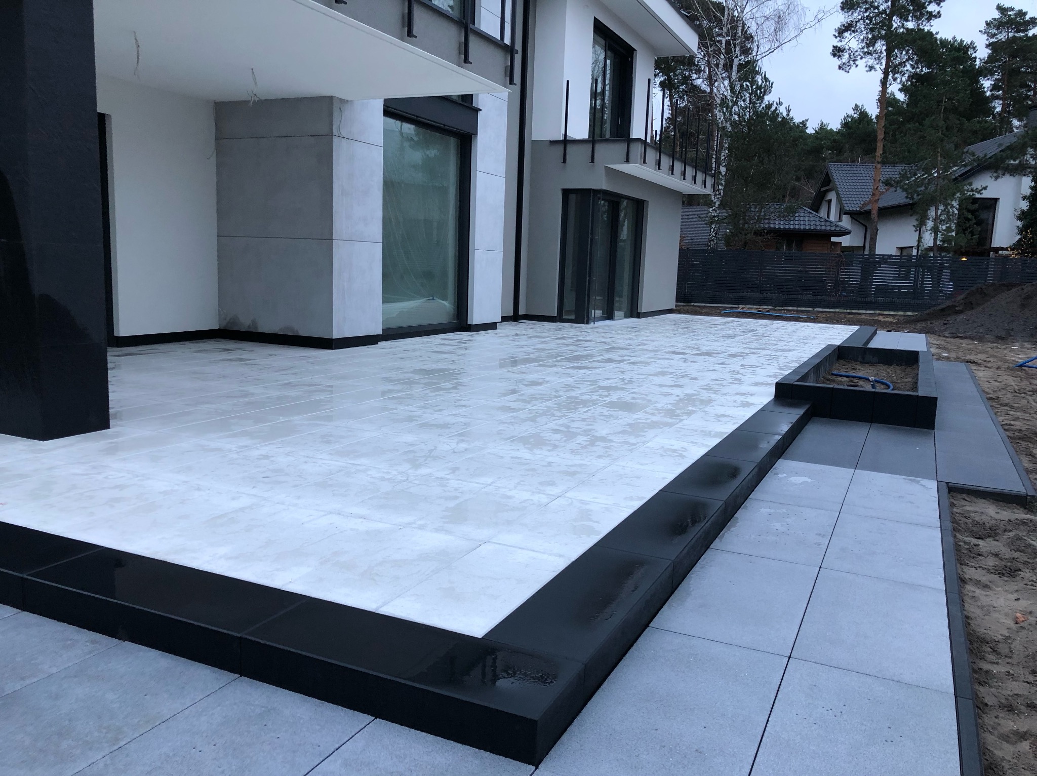 Nowoczesny taras z jasnoszarej kostki brukowej, obramowany czarnym, błyszczącym obrzeżem. Widok na dom z dużymi oknami i minimalistyczną architekturą.