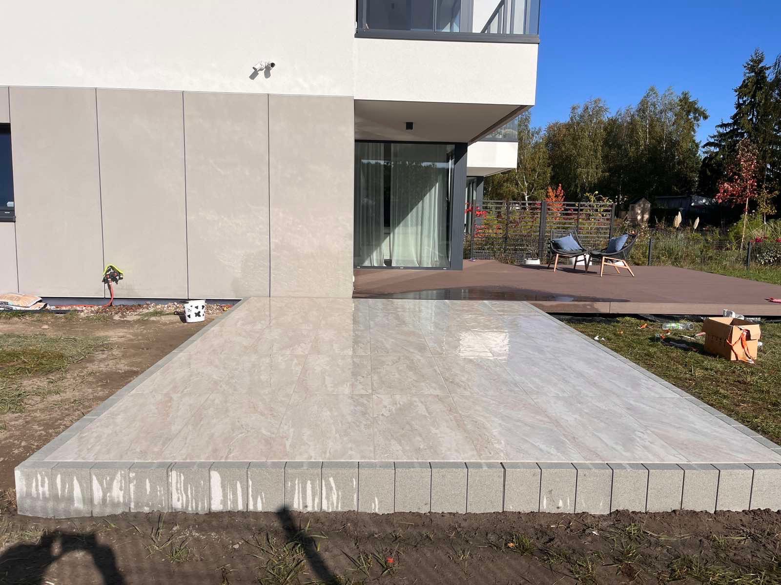 Wyłożony płytami taras z obrzeżem z kostki brukowej przy nowoczesnym domu z tarasem z desek kompozytowych i meblami ogrodowymi w tle.